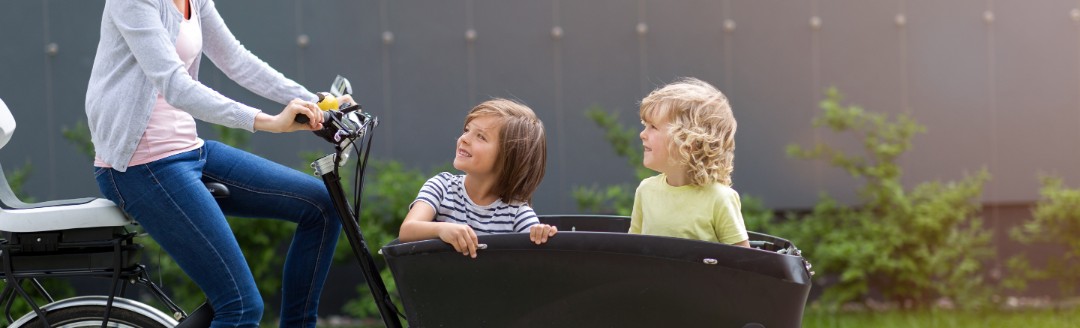 Eine Mutter fährt ein Lastenrad. Vorne im Anhänger sitzen zwei Kinder, die ihre Mutter anschauen. 