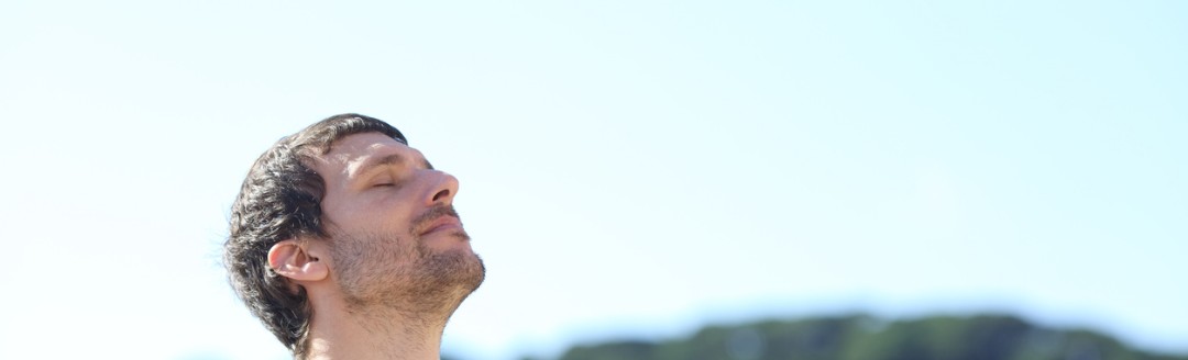 Ein Mann steht draußen mit geschlossenen Augen und hebt das Gesicht leicht in die Sonne, im Hintergrund eine grüne Hügellandschaft.