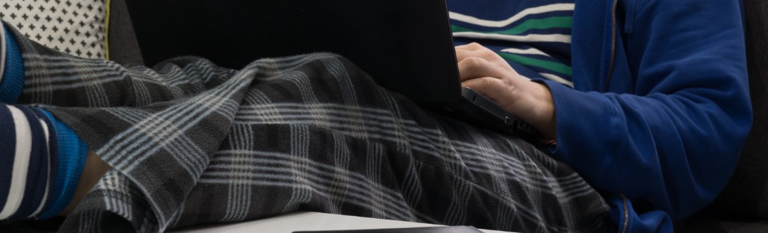 Eine männliche Person (Gesicht nicht zu sehen) liegt auf dem Sofa in Jogginghose und arbeitet am Laptop.