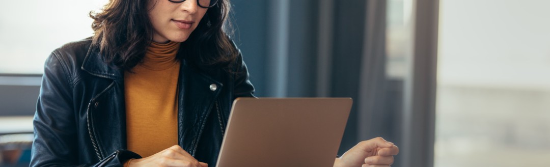 Eine Frau sitzt an einem Tisch und schaut auf ihren Laptop. Neben ihr liegen Dokumente. 