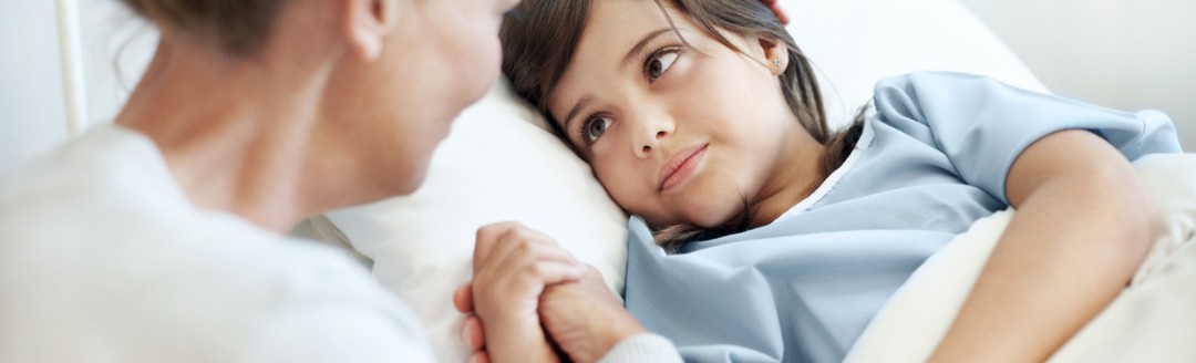 Ein kleiner Junge liegt im Krankenbett. Seine Mutter sitzt daneben und streichelt ihm den Kopf.