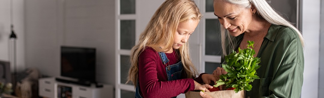 Eine ältere Frau schaut sich mit einem jüngeren Mädchen die Einkäufe in einer Papiertüte an. Sie sitzen zusammen in der Küche.