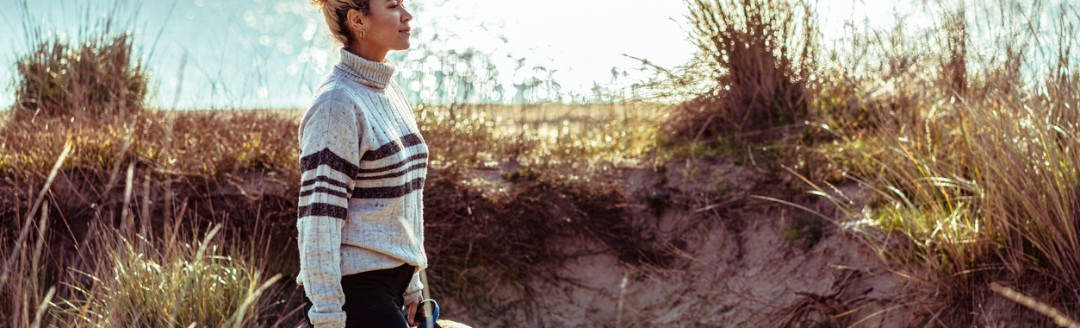 Eine Frau geht mit einem Hund an der Küste entlang, umgeben von Dünen und Meer im Hintergrund.