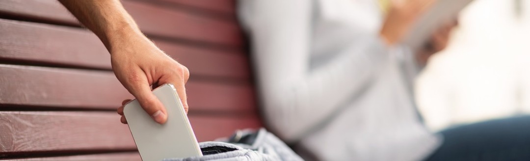 Eine Frau liest ein Buch auf einer Parkbank, während ihr Handy von einem Fremden aus der Tasche gezogen wird. 