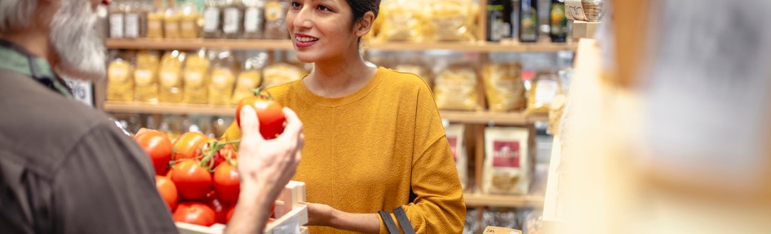 Eine junge Frau lächelt, während sie mit einem älteren Mann in einem Laden spricht und dabei einen Korb mit frischen Lebensmitteln hält. Im Hintergrund sind Regale mit verpackten Produkten zu sehen.
