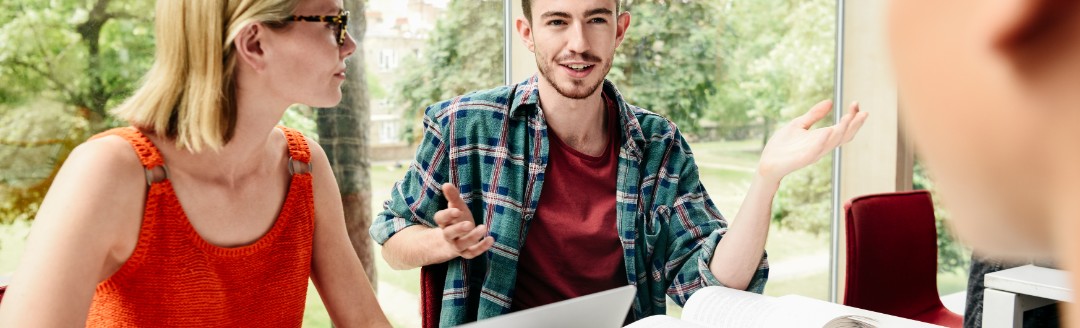 Eine Gruppe von jungen Leuten sitzen zusammen an einem Tisch mit Unterlagen und Laptops. Sie unterhalten sich. 