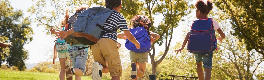 Mehrere Kinder laufen mit ihren Schulrucksäcken über eine Wiese in der Sonne.