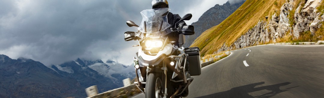 Motorradfahrer fährt auf einer kurvigen Bergstraße, im Hintergrund sind Berge und dunkle Wolken zu sehen.