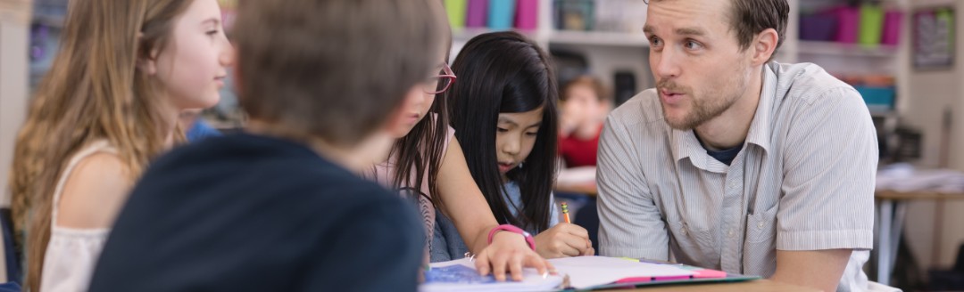 Ein junger Lehrer unterhält sich  mit zwei Schülern. Sie sitzen zu dritt an einem Tisch in einer Bibliothek. 