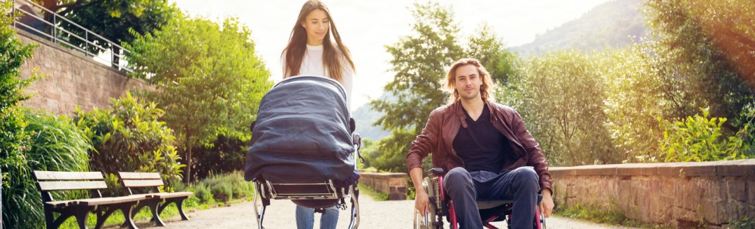 Eine Frau schiebt einen Kinderwagen, während ein Mann daneben in einem Rollstuhl sitzt. Sie spazieren gemeinsam einen Weg entlang. 