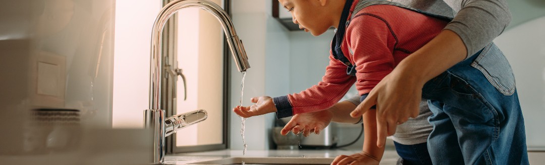 Kleiner Junge steigt auf einem Stuhl, um sich die Hände zu waschen während seine Mutter ihm hilft.