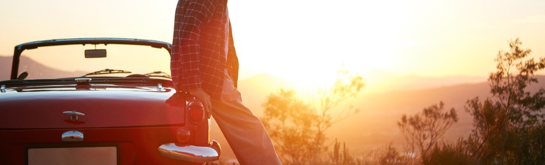 Ein erwachsener Mann lehnt an seinem Oldtimer und schaut von einem Hügel aus in den Sonnenuntergang. 