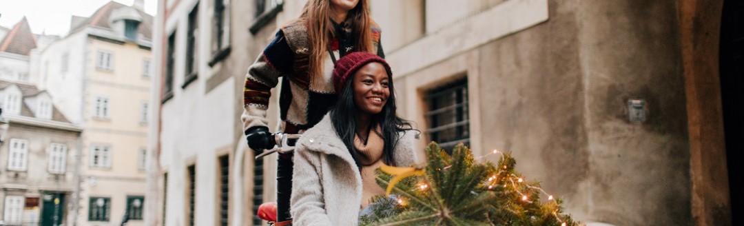 Zwei Freundinnen sind zusammen mit einem Lastenrad durch die Straßen unterwegs. Eine Person fährt das Rad, die andere Person sitzt drin. Sie transportieren einen kleinen Nadelbaum.