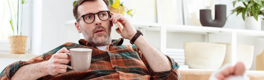 Ein Mann mit Brille sitzt auf einem Sofa, telefoniert und hält eine Tasse. Sein Bein ist in Gips und hochgelegt.