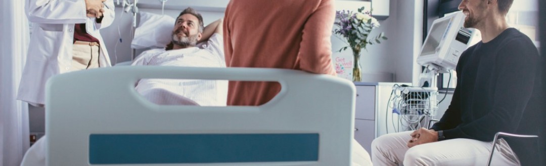 Ein männlicher Patient liegt in einem Krankenhausbett. Rechts von ihm steht eine weibliche Ärztin, eine Frau sitzt am Fußende des Bettes und ein Mann daneben auf einem Stuhl. 