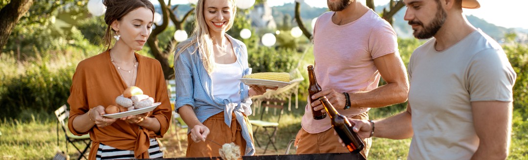 Eine Gruppe von Freunden steht zu viert an einem Holzgrill im Grünen. 