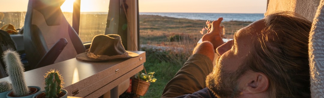 Ein junger Mann liegt in seinem ausgebauten Campervan mit Sonne im Hintergrund.