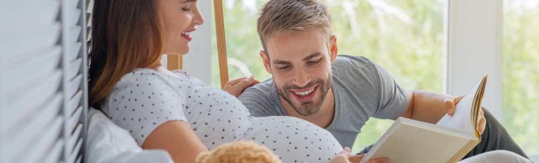 Eine schwangere Frau liegt im Bett. Ein Mann liegt neben ihr und liest ein Bett vor, während er auf den Babybauch schaut. 
