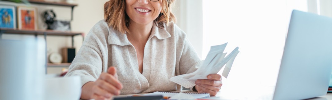 Frau mit Brille sitzt lächelnd an einem Schreibtisch, hält Belege in der Hand und arbeitet mit Taschenrechner und Laptop.
