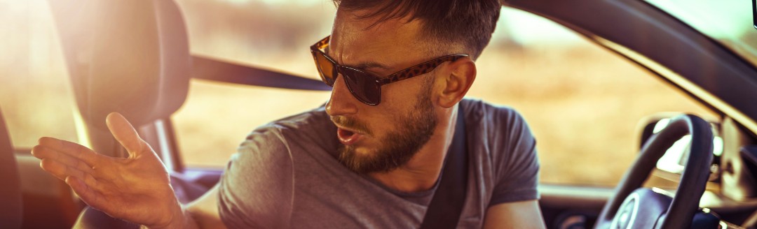 Ein Mann mit Sonnenbrille schaut verwundert und Schultern zuckend um sich im Auto.  