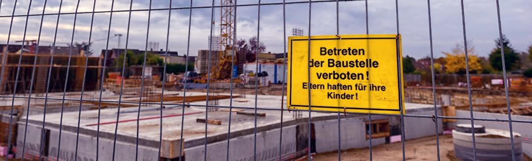 Das Bild zeigt eine Baustelle, welche mit einem Zaun abgesperrt ist. An dem Zaun hängt ein gelbes Warnschild, auf dem "Betreten der Baustelle verboten! Eltern haften für ihre Kinder!" steht. 