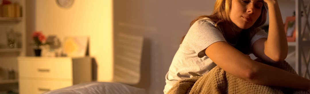 Eine Frau sitzt krank und erschöpft in ihrem Bett. Sie hält sich ein Wärmekissen an die Stirn. 