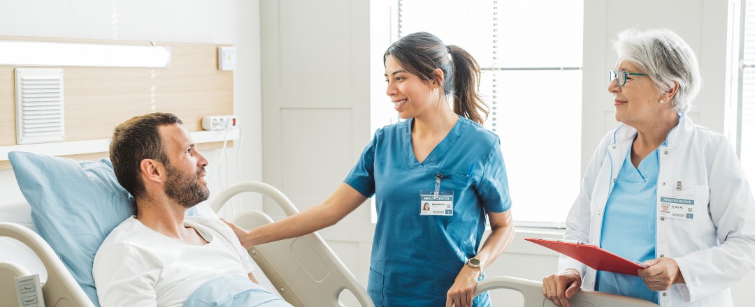 Eine junge Pflegerin und eine ältere Ärztin stehen gemeinsam an einem Patientenbett und unterhalten sich mit dem dort liegenden männlichen Patienten.