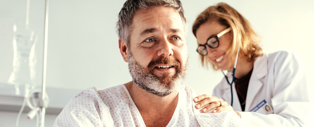 Eine Ärztin hört mit einem Stethoskop den Rücken eines männlichen Patienten ab, welcher aufrecht im Patientenbett sitzt.