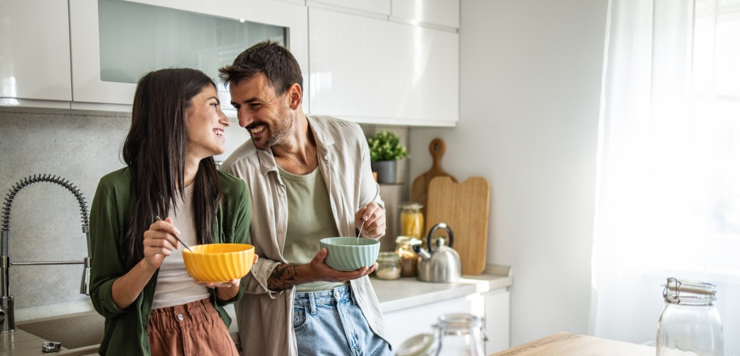 Ein junges Paar steht mit Müslischalen in der Hand steht lachend in der Küche