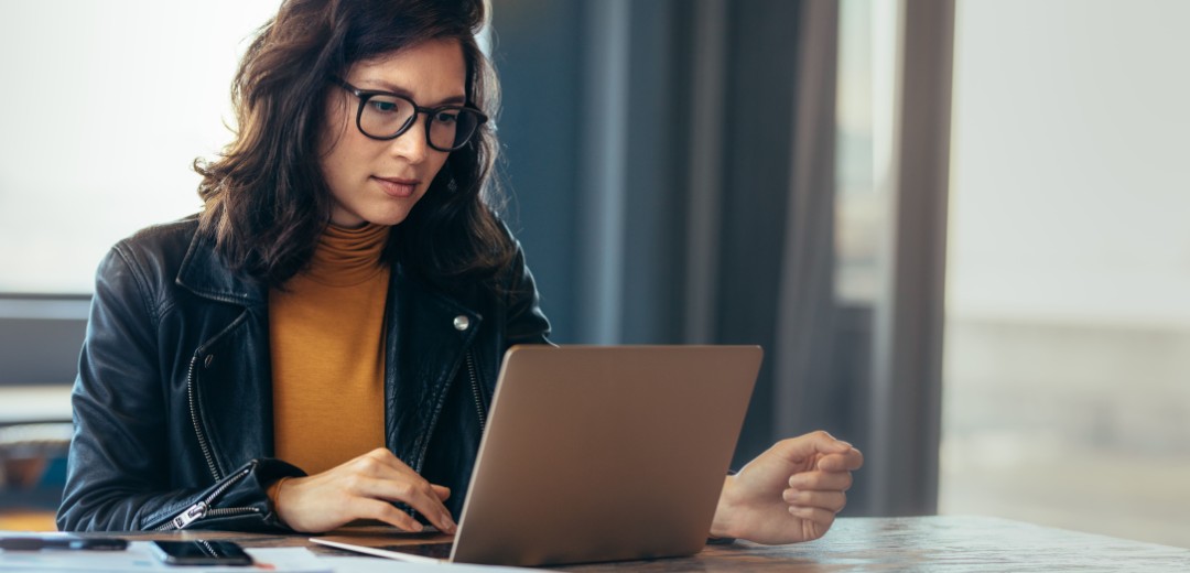 Eine Frau sitzt an einem Tisch und schaut auf ihren Laptop. Neben ihr liegen Dokumente. 