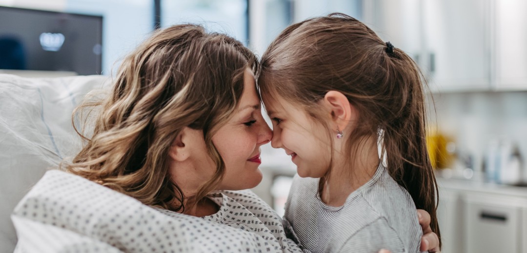 Eine Frau im Krankenbett und ihre kleine Tochter geben sich einen Nasenkuss