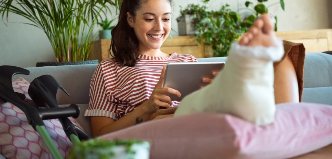 Fröhliche junge Frau sitzt mit gebrochenem Bein zu Hause.