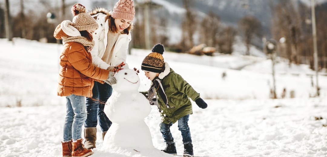 Eine Mutter baut mit ihren zwei Kindern einen Schneemann in einer Winterlandschaft. 