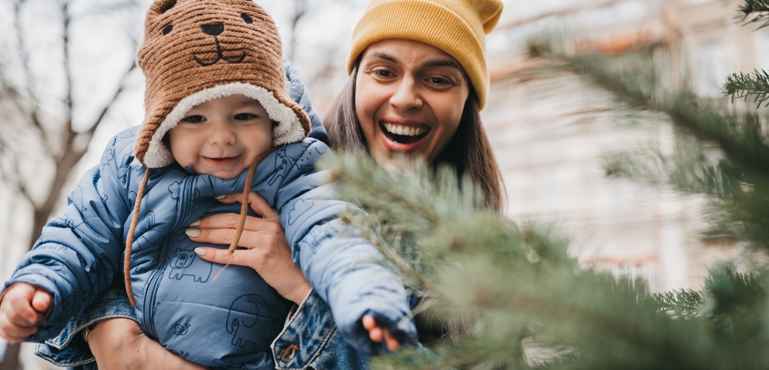 Glückliche Frau hält ihr Baby vor einem Tannenbaum.
