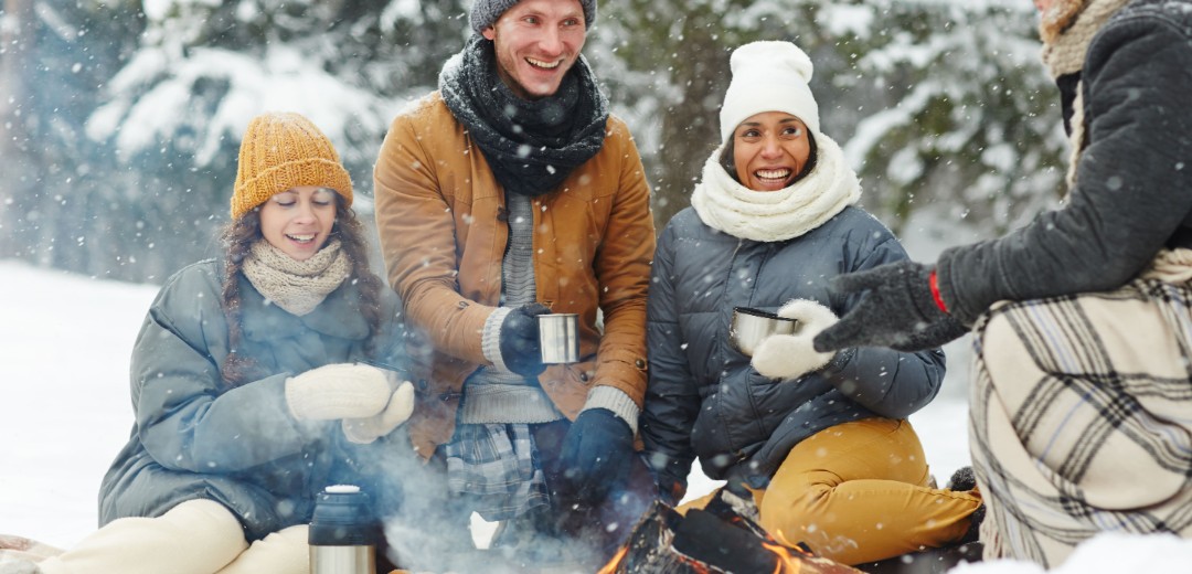 Eine Gruppe von Freunden sitzt in Winterklamotten im Schnee und grillt. 
