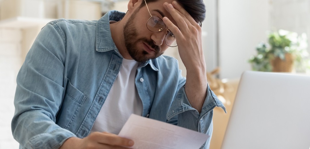 Ein Mann sitzt am Laptop und schaut frustriert auf ein kleineres Blatt Papier. 