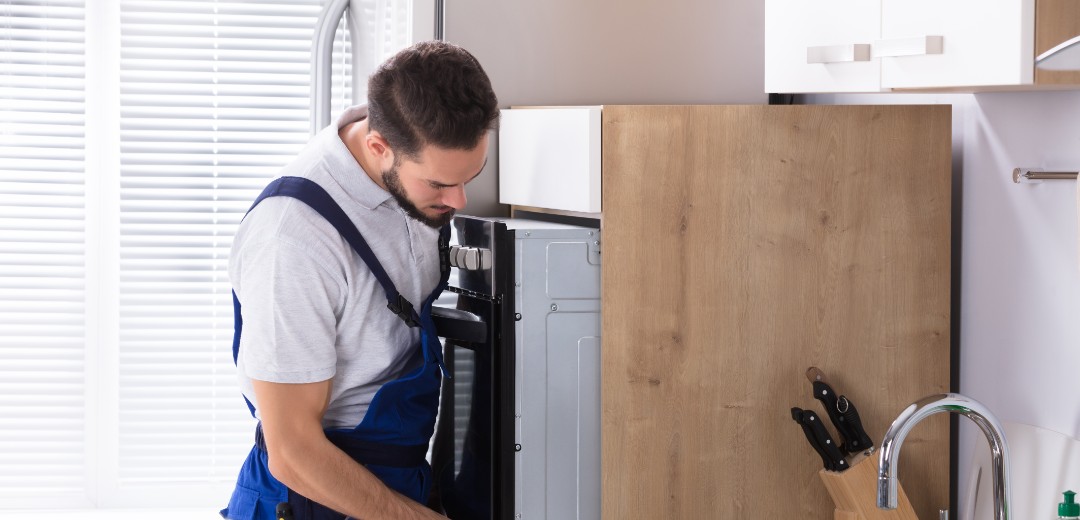 Ein Elektriker steht in einer Küche und untersucht einen Ofen, den er zuvor leicht aus dem Küchenschrank gezogen hat.