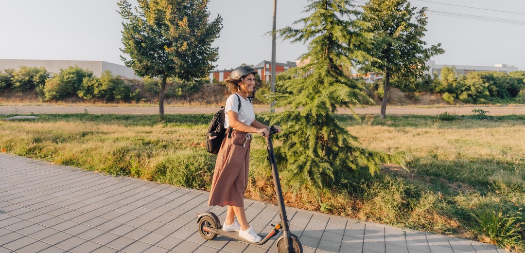 Junge Frau mit Helm fährt auf einem E-Scooter durch eine Grünanlage.