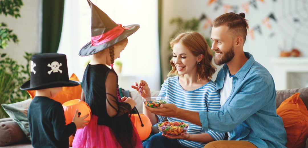 Zwei auf dem Sofa sitzende Eltern geben ihren verkleideten Kindern Süßigkeiten in ihren Halloween-Eimer. 
