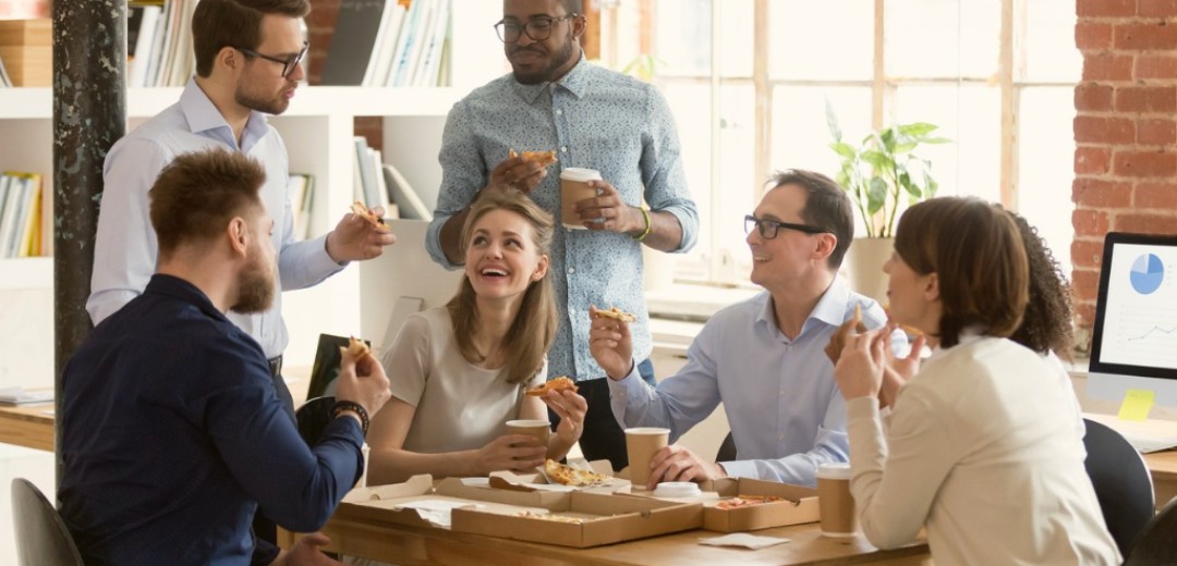 Eine Gruppe von Kollegen sitzt lachend zusammen im Büro an einem Tisch und isst Pizza. 