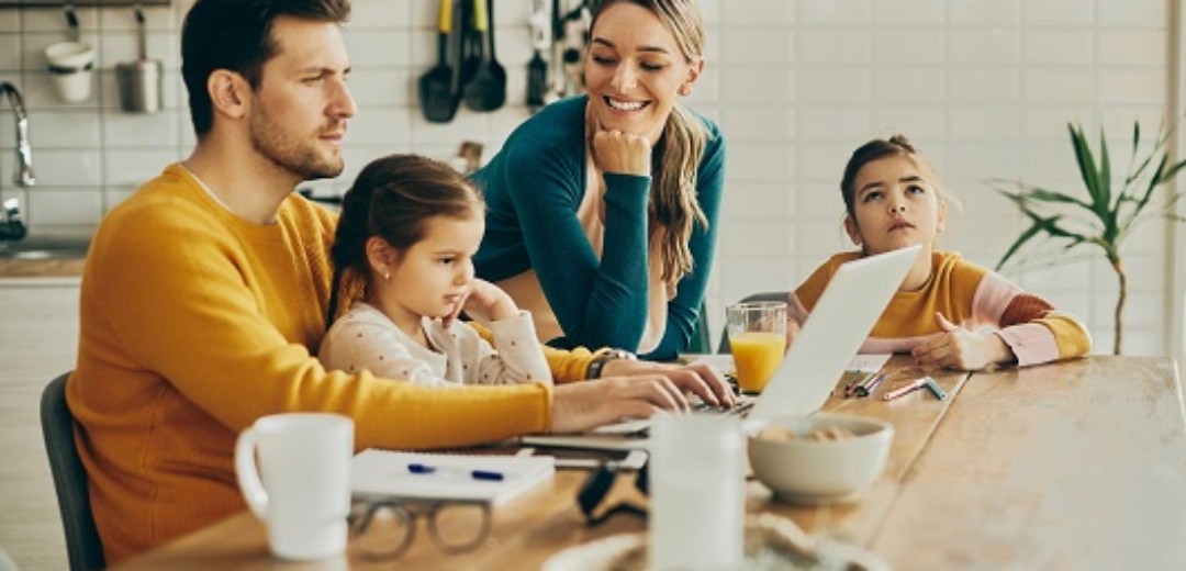 Eine junge Familie sitzt zusammen am Küchentisch. Der Vater arbeitet an seinem Laptop.