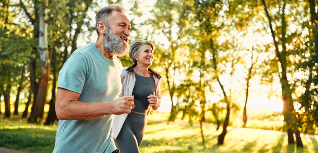 Ein älteres Paar joggt gemeinsam lachend durch einen sonnigen Park.