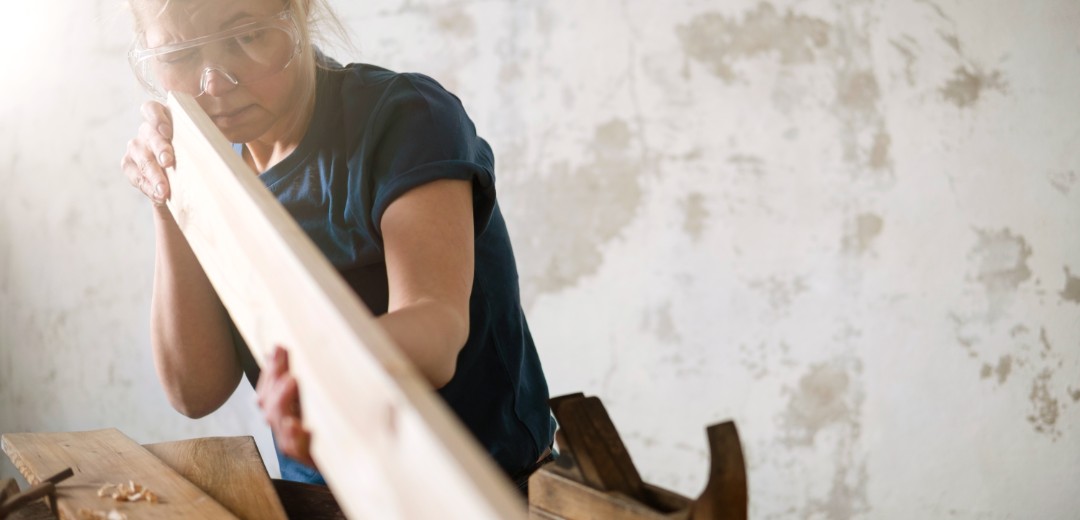 Eine Frau arbeitet an einem länglichen Stück Holz. Sie trägt eine Schutzbrille und neben ihr liegen diverse Werkzeuge. 