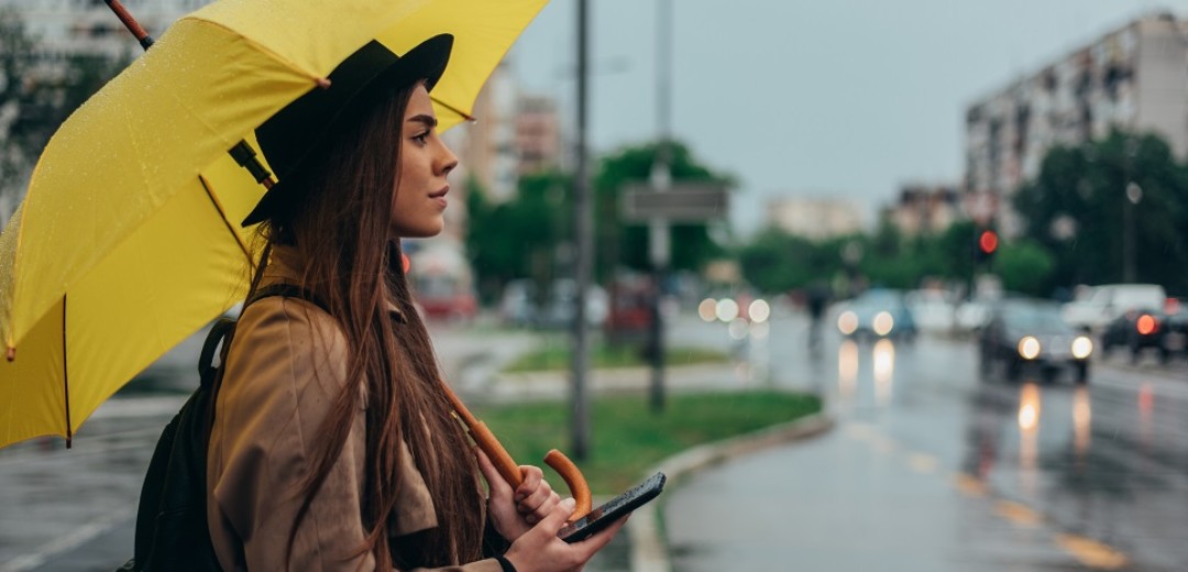 Eine junge Frau steht im Regen mit einem gelben Regenschirm am Straßenrand und sucht mit ihrem Handy in der Hand etwas. 