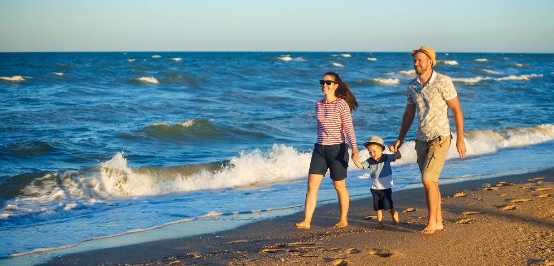 Ein Pärchen läuft mit ihrem Kleinkind Hand in Hand einen Strand entlang. 
