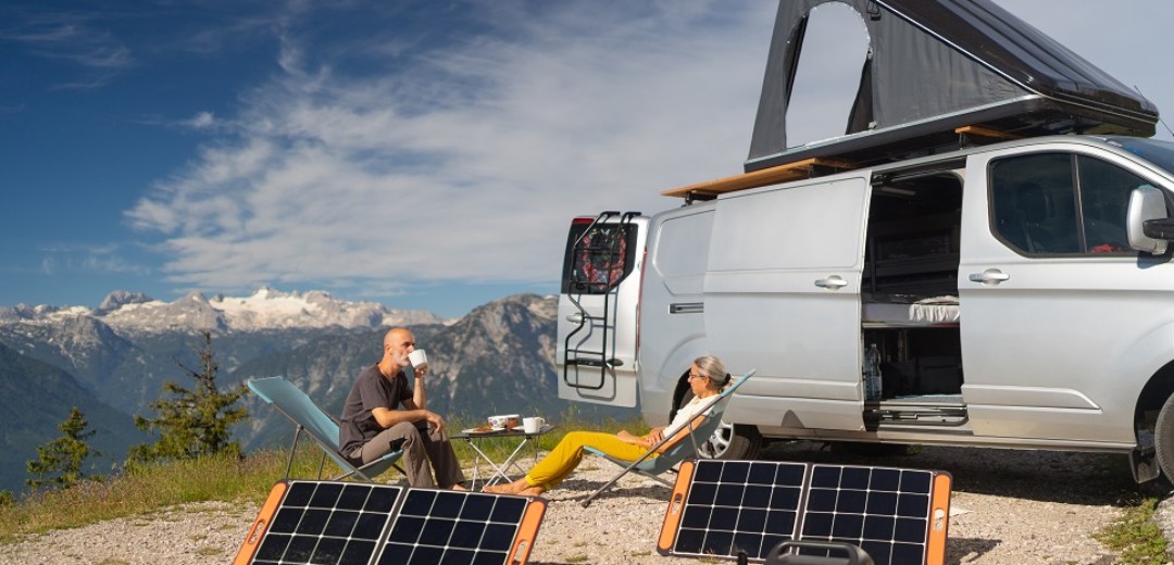 Eine Frau und ein Mann sitzen zusammen in einem Liegestuhl vor ihrem Campervan in den Bergen. Vor ihnen stehen zwei Solarpanels.