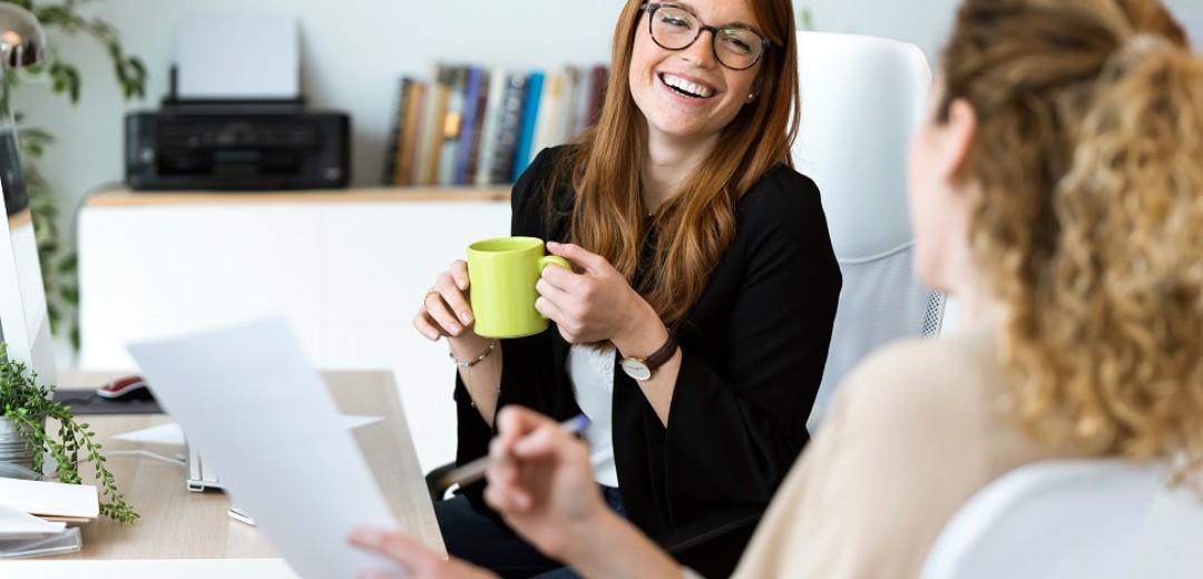 Zwei junge Frauen unterhalten sich lachend am Arbeitsplatz.