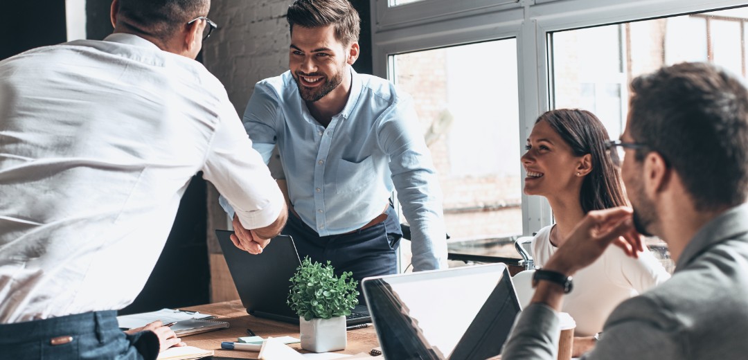 Eine kleinere Gruppe von Leuten sitzen zusammen im Büro und zwei Personen reichen sich die Hände. Sie tragen alle Business-Kleidung. 