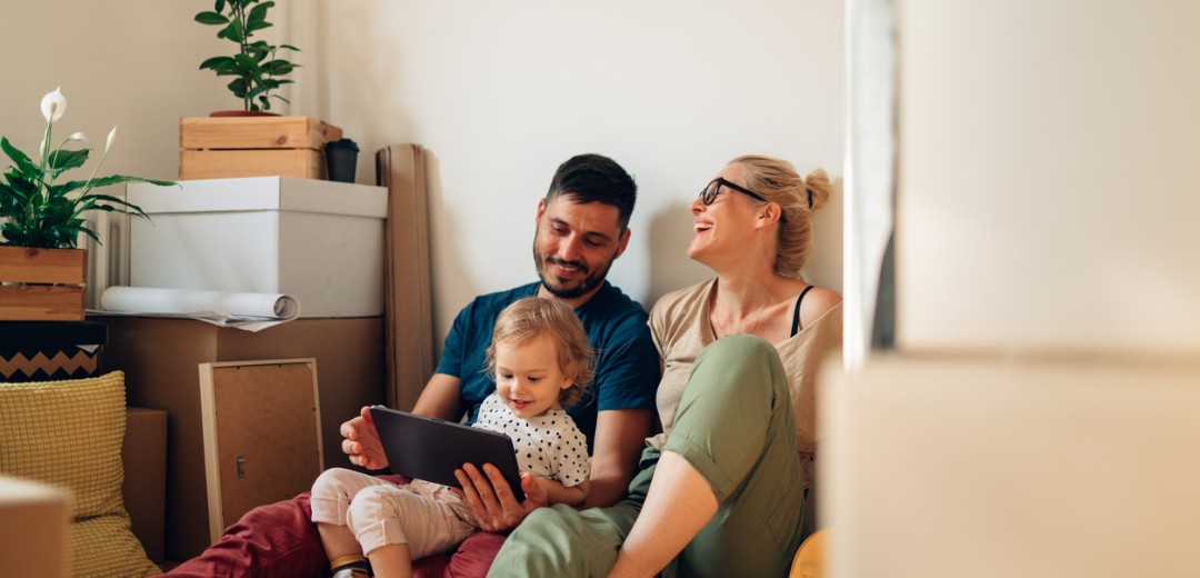 Eine Familie sitzt zwischen Umzugskartons auf dem Boden; ein Mann und ein kleines Kind schauen gemeinsam auf ein Tablet, während eine Frau daneben lachend zusieht.