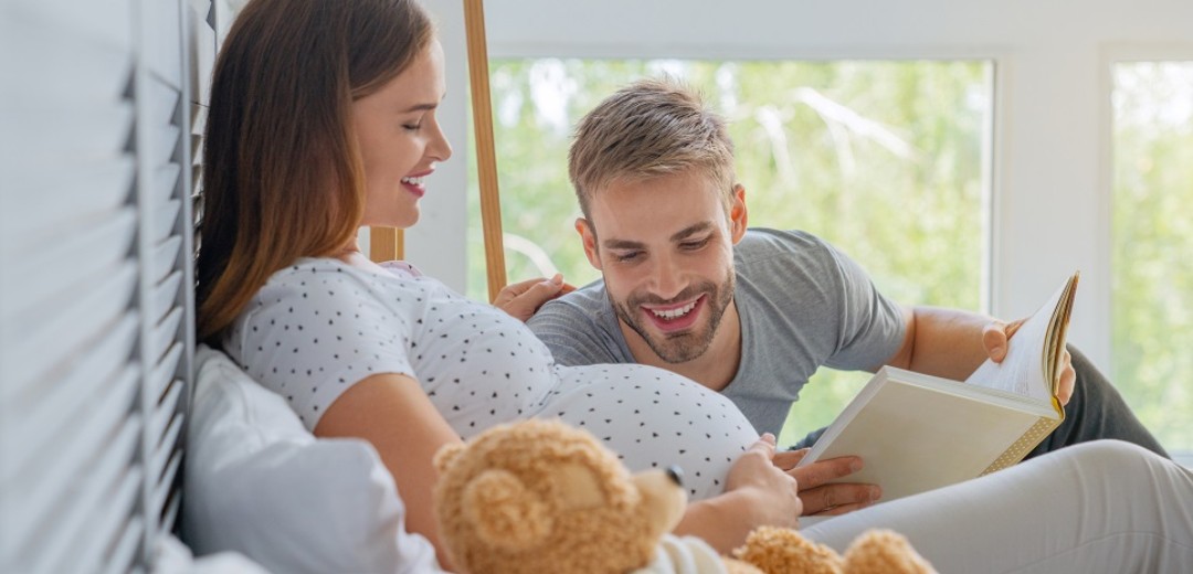Eine hochschwangere Frau liegt aufrecht im Bett, neben ihr ein Teddybär und ein Mann liest ein Buch vor, während er auf den Babybauch schaut. 